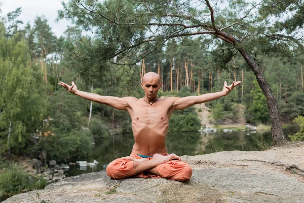 Sem camisa budista meditando em pose de lótus com as mãos estendidas em penhasco rochoso sobre o lago — Fotografia de Stock