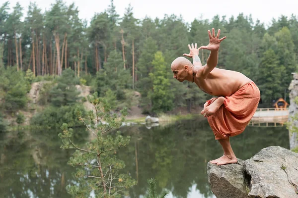 Shirtless buddhist meditating in yoga pose with outstretched hands on rocky cliff over river — Stock Photo