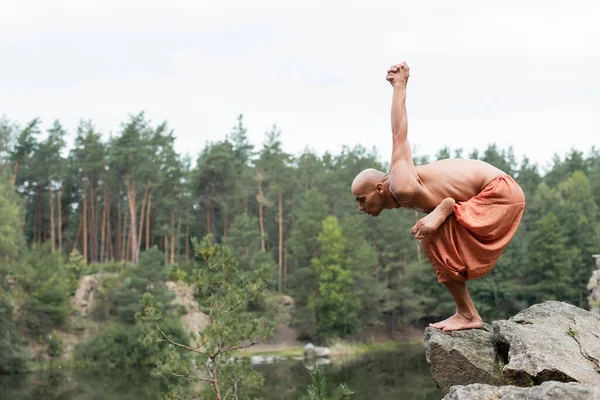 Side view of shirtless buddhist meditating in yoga pose on rocky cliff over forest lake — Stock Photo