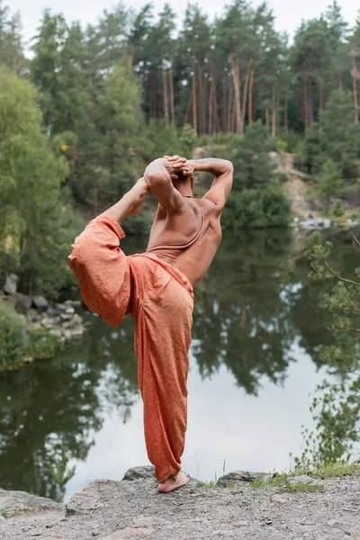Shirtless buddhist in harem pants practicing one legged bow pose on rocky cliff over forest lake — Stock Photo