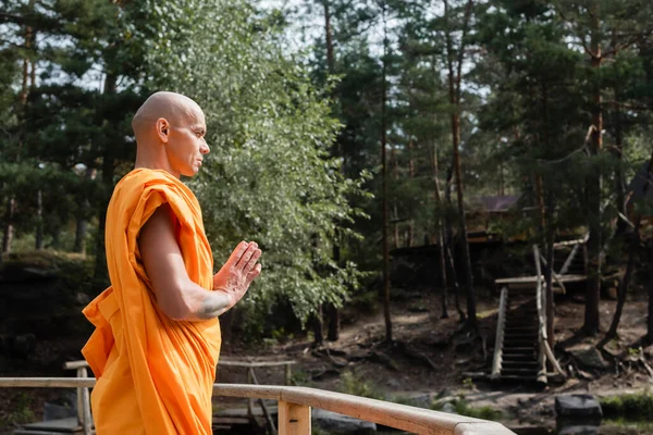 Side view of buddhist meditating with praying hands near wooden fence in forest — Stock Photo