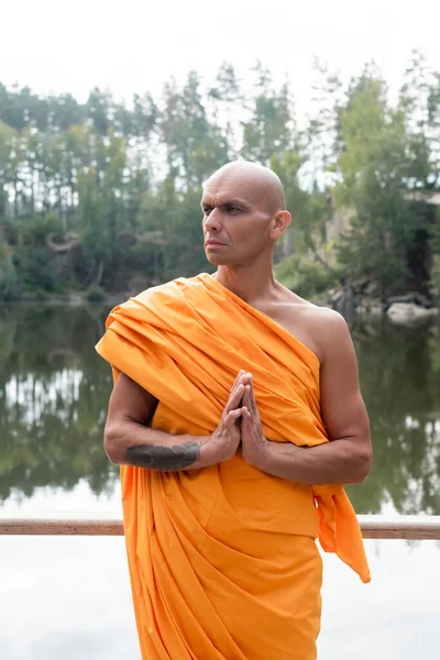 Buddhist monk in traditional robe looking away while meditating near lake in forest — Stock Photo