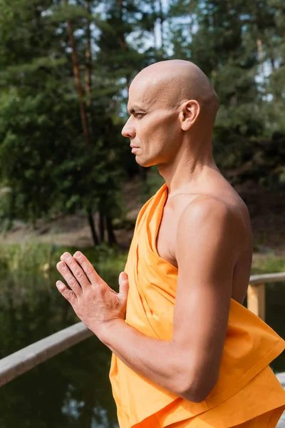 Vista lateral de budista em roupão laranja meditando com as mãos de oração ao ar livre — Fotografia de Stock
