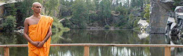 Buddhist in traditional robe looking away near wooden fence over lake in forest, banner — Stock Photo