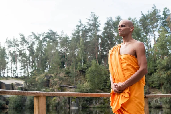 Буддийский монах в оранжевой касайе смотрит в сторону, медитируя возле деревянного забора в лесу — стоковое фото