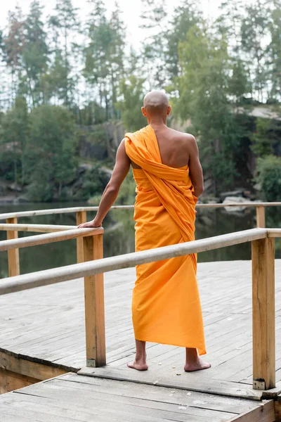 Vista trasera del hombre descalzo en búdhist bata meditando en plataforma de madera cerca del lago - foto de stock