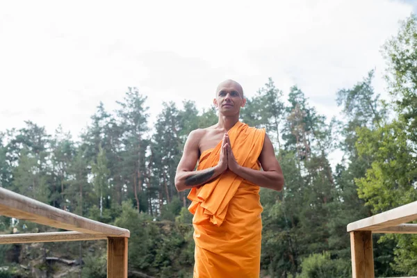 Low angle view of buddhist monk meditating with praying hands near wooden fence in forest — Stock Photo