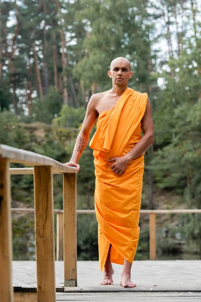 Vista completa del monje budista de pie cerca de una cerca de madera en el bosque - foto de stock