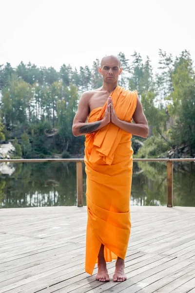 Vista completa del monje budista meditando con las manos orantes en la plataforma de madera cerca del lago del bosque - foto de stock