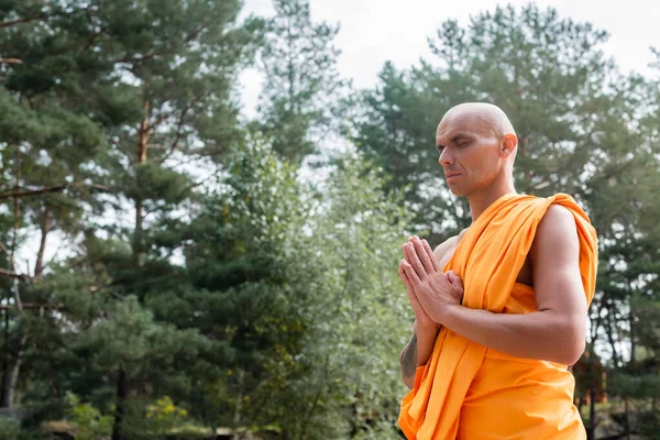 Buddhist en bata naranja rezando con los ojos cerrados al aire libre - foto de stock