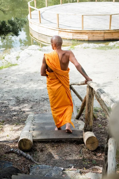 Rückansicht eines haarlosen Mannes in traditioneller buddhistischer Kleidung, der auf Holztreppen im Wald wandelt — Stockfoto