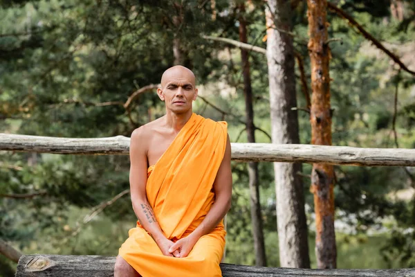 Buddhist sentado en el banco de troncos y meditando con los ojos cerrados en el bosque - foto de stock