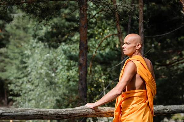 Homem em budista roupas tradicionais de pé perto de cerca de madeira na floresta e olhando para longe — Fotografia de Stock