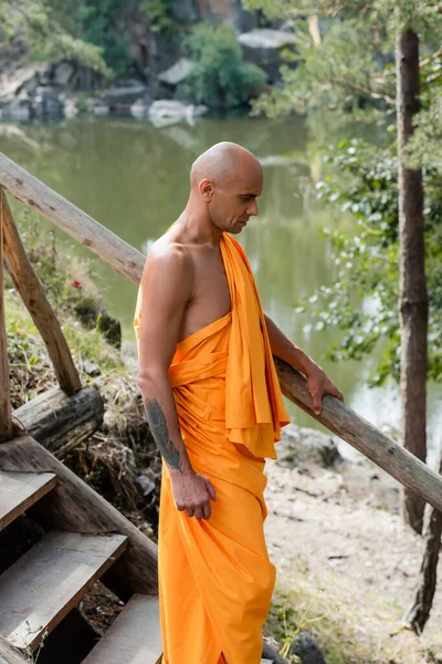 Buddhist monge em roupão laranja andando em escadas de madeira perto do lago — Fotografia de Stock