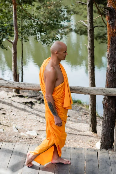 Side view of buddhist monk walking on wooden walkway near forest lake — Stock Photo