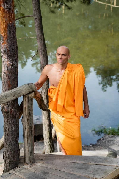 Buddhist monk in traditional orange robe walking on wooden stairs near pond — Stock Photo