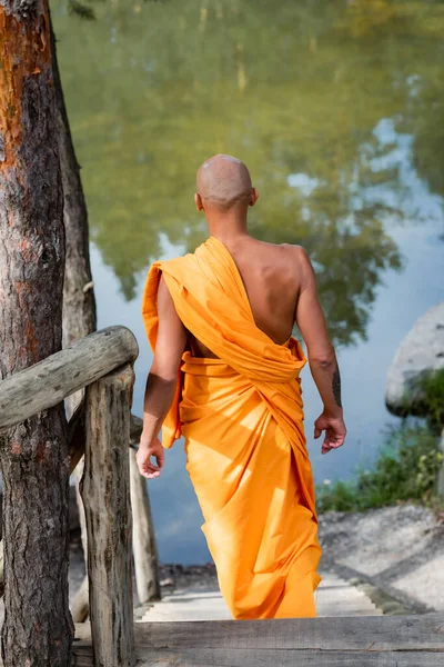 Vista trasera del monje budista caminando por las escaleras en el bosque cerca del lago - foto de stock