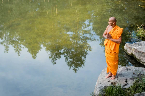 High angle view of buddhist in orange kasaya meditating with praying hands near lake — Stock Photo