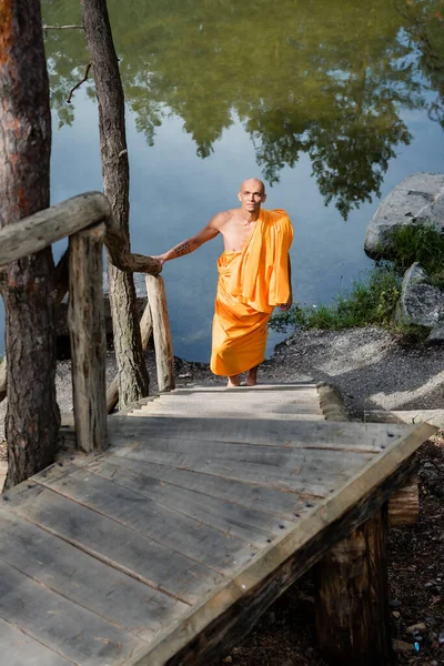 High angle view of buddhist in orange robe walking on wooden stairs near lake — Stock Photo