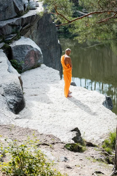 Vista de ángulo alto del monje budista meditando cerca de rocas sobre el lago del bosque - foto de stock