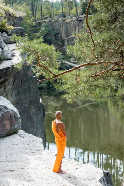 High angle view of buddhist in orange kasaya meditating on rocky cliff over water — Stock Photo