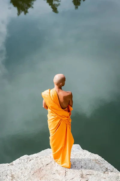 Vista de ángulo alto de budista en kasaya naranja meditando en acantilado rocoso sobre el lago - foto de stock