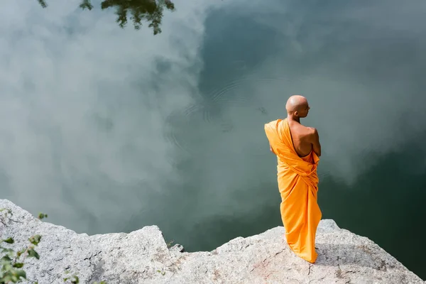Visão de alto ângulo do monge budista meditando em penhasco rochoso sobre a água — Fotografia de Stock