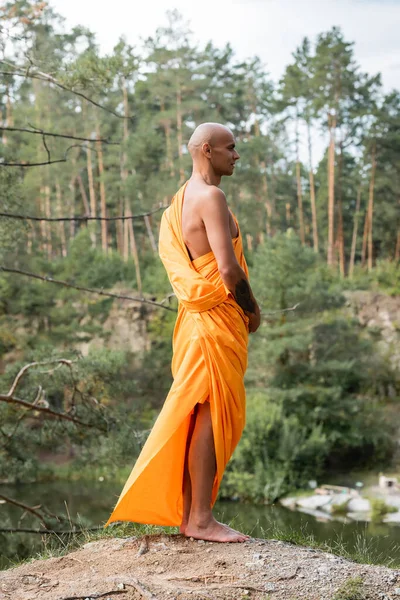 Tattooed buddhist in traditional orange robe meditating on hill in forest — Stock Photo