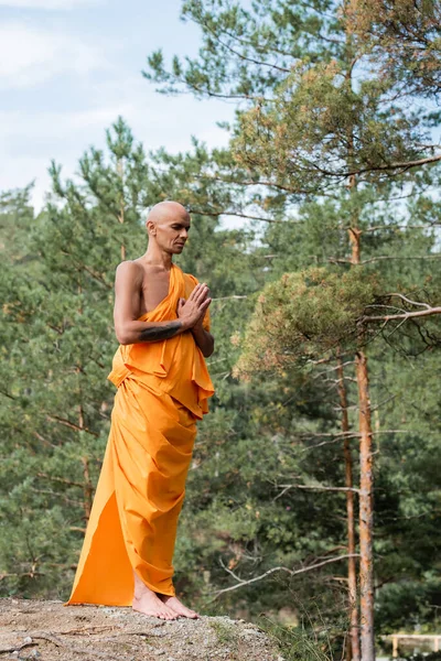 Ganzkörperansicht des Buddhisten auf orangefarbenem Kasaya, der mit geschlossenen Augen im Wald betet — Stockfoto