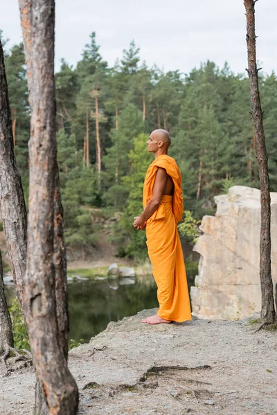 Vista completa del monje budista meditando sobre un acantilado rocoso en el bosque - foto de stock
