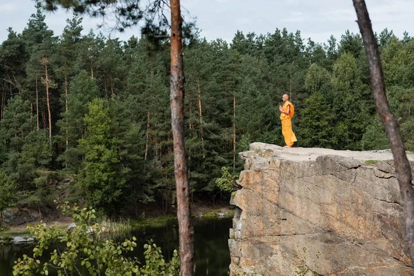 Monje budista en kasaya naranja rezando en alto acantilado rocoso sobre el río en el bosque - foto de stock