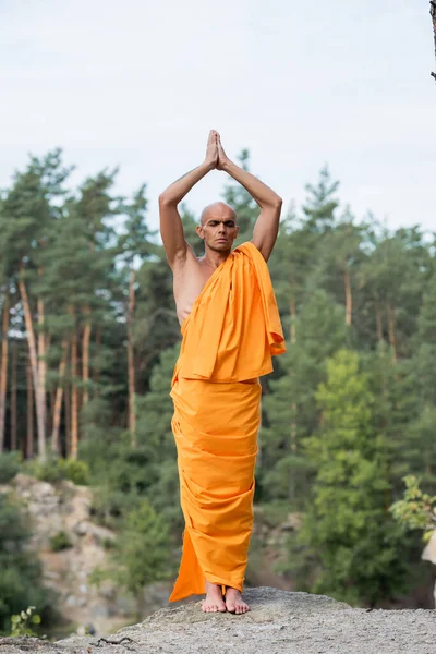 Vista completa de budista en kasaya naranja meditando con las manos orantes levantadas en el bosque - foto de stock