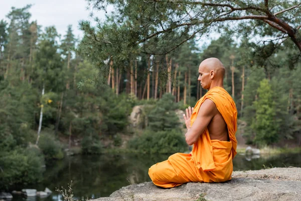 Monje budista en kasaya naranja meditando en pose de loto con manos orantes cerca del lago del bosque - foto de stock
