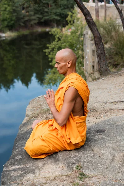 Буддист в оранжевом халате медитирует в позе лотоса с молящимися руками на скалистой скале возле воды — стоковое фото