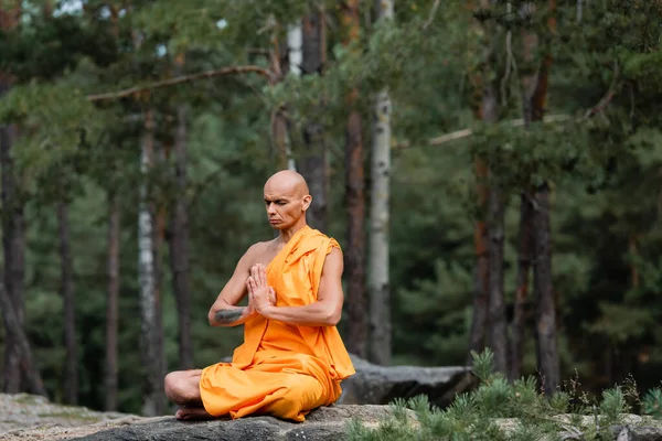 Buddhist monk in orange kasaya sitting in lotus pose with praying hands while meditating in forest — Stock Photo