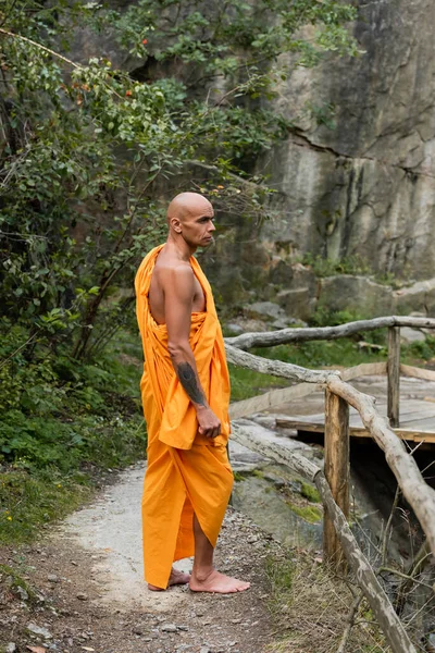 Visão de comprimento total do budista descalço em roupão laranja em pé perto de cerca de madeira na floresta — Fotografia de Stock