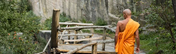 Vista posteriore dell'uomo in vestaglia buddista passeggiando su passerella in legno nella foresta, banner — Foto stock