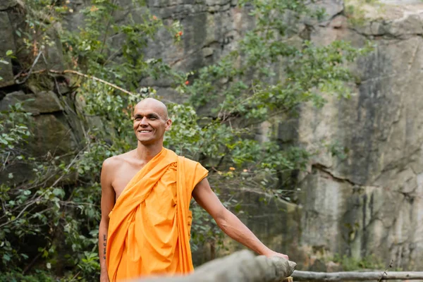 Happy buddhist in orange kasaya walking near wooden fence in forest — Stock Photo