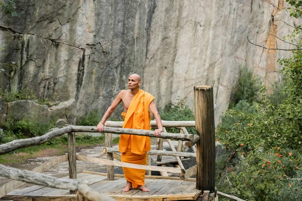 Buddhist in orange kasaya standing on wooden walkway near rocks and looking away — Stock Photo