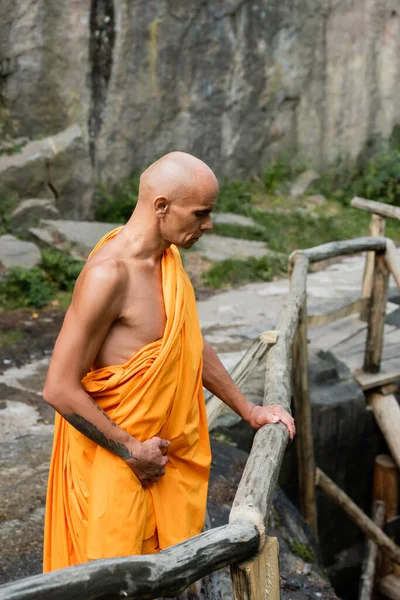 Tattooed buddhist in traditional robe looking down while standing near wooden fence — Stock Photo