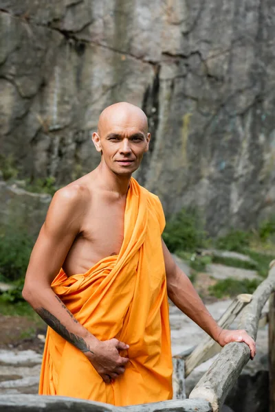 Buddhist en kasaya naranja mirando la cámara mientras está parado cerca de la cerca de madera al aire libre - foto de stock