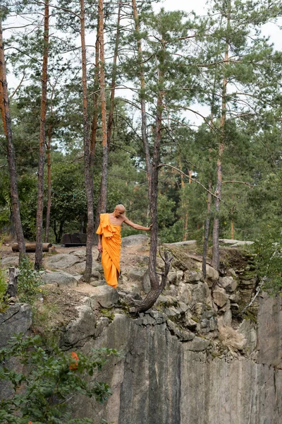 Повний вигляд на буддиста в помаранчевому халаті, що йде по скелястій скелі в лісі — стокове фото