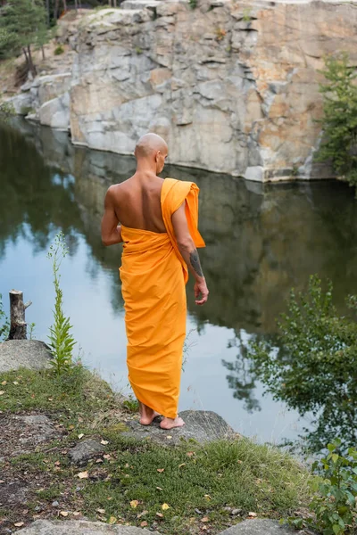 Back view of barefoot buddhist in orange robe meditating near lake in forest — Stock Photo