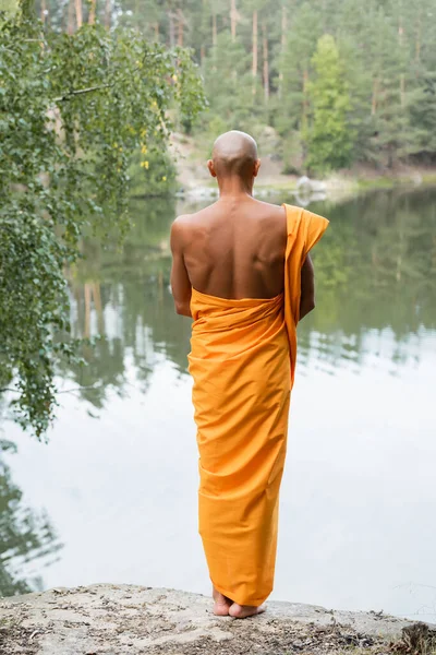 Back view of bald man in traditional buddhist robe meditating near forest pond — Stock Photo