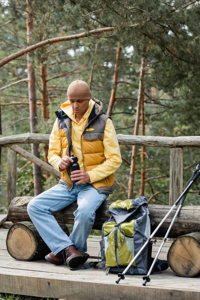 Turista em garrafa térmica de abertura de colete quente durante parada no banco de log perto de mochila e postes de trekking — Fotografia de Stock