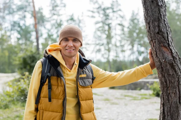 Heureux voyageur en bonnet et gilet chaud souriant à la caméra dans la forêt — Photo de stock