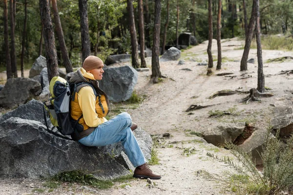 Excursionista con mochila sentada en piedra en el bosque y mirando hacia otro lado - foto de stock