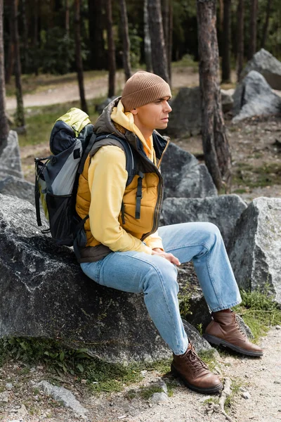 Traveler with backpack, wearing warm vest and beanie, sitting on stone in forest — Stock Photo