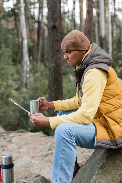 Viajante em gorro e colete quente sentado com caneca de metal e mapa na floresta — Fotografia de Stock