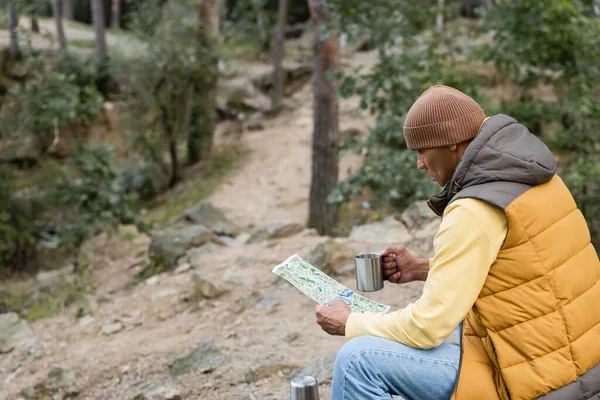 Randonneur en bonnet et gilet chaud assis dans la forêt avec carte et tasse en métal — Photo de stock